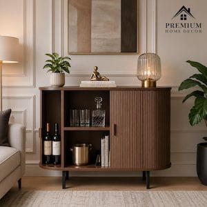 May include: A dark brown wooden sideboard with a curved tambour door, open shelving, and black legs. The unit holds two wine bottles, three clear glasses, a glass decanter, a metal ice bucket, and two books. The logo Premium Home Decor is visible.