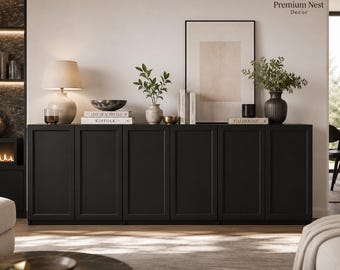 May include: A long, matte black sideboard cabinet with six rectangular panel doors. The top surface holds a beige table lamp, a dark bowl, a small vase with green foliage, several stacked books, and a framed abstract print on the wall.