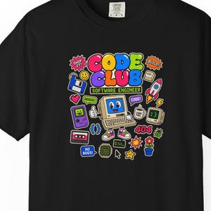 以下が含まれることがあります： 「CODE CLUB SOFTWARE ENGINEER」というテキストがプリントされたカラフルなグラフィックデザインの黒いTシャツ。デザインには、漫画風のコンピューターキャラクター、ロケット、フロッピーディスク、カセットテープ、その他様々な小さなテクノロジー関連のアイコンが含まれています。