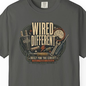 May include: A charcoal gray short-sleeve t-shirt featuring a graphic print of electrical tools, a gauge, and a power plug. The text reads WIRED DIFFERENT, BUILT FOR THE CIRCUIT, TEST, TRACE, REPAIR, and ELECTRICAL ENGINEERING FIELD UNIT.