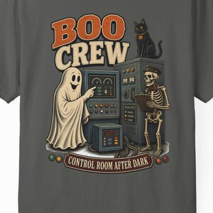 Może przedstawiać: Ciemnoszary t-shirt z grafiką przedstawiającą ducha, szkielet i czarnego kota w sterowni. Napis głosi BOO CREW oraz CONTROL ROOM AFTER DARK. Wzór zawiera elementy maszyn przemysłowych z pokrętłami, przyciskami i wskaźnikami pomiarowymi.