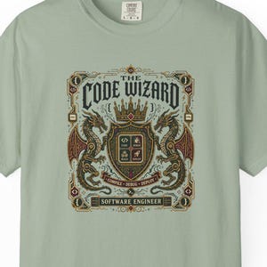 Op de afbeelding: Een saliegroen T-shirt met korte mouwen en een centrale afbeelding van twee draken die een schild met een kroon flankeren. De tekst luidt THE CODE WIZARD, COMPILE DEBUG DEPLOY en SOFTWARE ENGINEER, met codesymbolen in het schild.