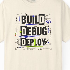 Puede incluir: Una camiseta de manga corta color crema con un gráfico central que muestra las palabras BUILD, DEBUG y DEPLOY en letras negras. El diseño incluye detalles en azul y verde lima, patrones de circuitos y símbolos de programación.