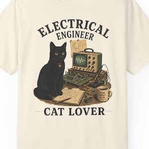 Puede incluir: Una camiseta de manga corta color hueso con un gráfico de un gato negro sentado en un escritorio con equipo electrónico, una placa de circuito y una taza. El texto ELECTRICAL ENGINEER y CAT LOVER aparece en letras oscuras sobre el diseño.