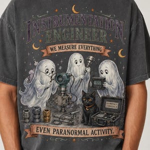 May include: A charcoal gray short-sleeve t-shirt featuring a graphic of three ghosts and a black cat using engineering tools. The text reads "INSTRUMENTATION ENGINEER WE MEASURE EVERYTHING EVEN PARANORMAL ACTIVITY." The design includes stars and moons.