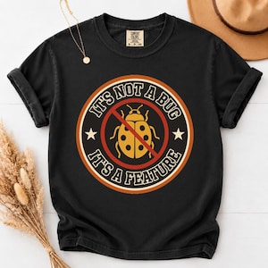 May include: A black short-sleeve t-shirt featuring a circular graphic with a yellow ladybug in the center, crossed out by a red line. The text reads IT'S NOT A BUG IT'S A FEATURE in white capital letters, surrounded by a rust-colored border.