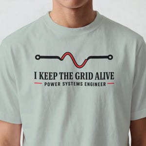 Könnte beinhalten: Ein helles salbeigrünes T-Shirt mit einer Grafik einer schwarzen und roten Stromkreislinie. Unter der Grafik steht der Text I KEEP THE GRID ALIVE in Schwarz, gefolgt von POWER SYSTEMS ENGINEER in Schwarz mit roten Linien.