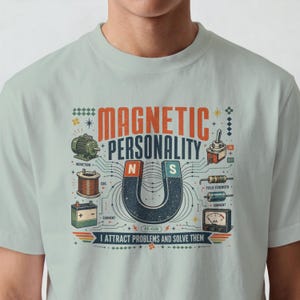 May include: A light sage green t-shirt featuring a graphic print with the text "MAGNETIC PERSONALITY" and "I ATTRACT PROBLEMS AND SOLVE THEM". The design includes a horseshoe magnet, an induction motor, a coil, a battery, and electrical meters.