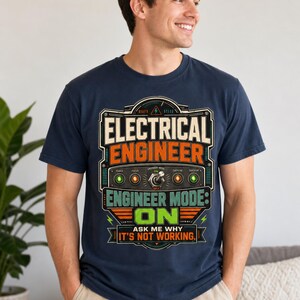 May include: A navy blue t-shirt featuring a graphic print of a control panel with the text ELECTRICAL ENGINEER, ENGINEER MODE: ON, and ASK ME WHY IT'S NOT WORKING. The design includes a central toggle switch, indicator lights, and gauges.