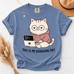 Könnte beinhalten: Ein blaues Kurzarm-T-Shirt mit einer Grafik einer weißen Katze, die eine runde Brille und einen rosa Kapuzenpullover trägt und an einem Laptop arbeitet. Auf dem Laptop ist ein Pfotenabdruck. Darunter steht der Text THIS IS MY DEBUGGING FACE.