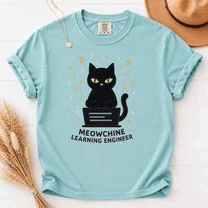 Puede incluir: Una camiseta azul claro con un gráfico de un gato negro sentado frente a un portátil, rodeado de dígitos binarios amarillos. Debajo del gato, el texto MEOWCHINE LEARNING ENGINEER aparece escrito en letras mayúsculas de color negro.