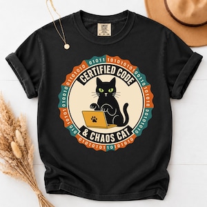 May include: A black short-sleeve t-shirt featuring a circular graphic with a black cat sitting at a laptop. The text reads CERTIFIED CODE & CHAOS CAT, surrounded by a border of binary code in orange, teal, and cream colors on a black background.