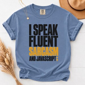 May include: A blue short-sleeved t-shirt with black and yellow text that reads I SPEAK FLUENT SARCASM AND JAVASCRIPT followed by a semicolon. The shirt features a crew neckline and a Comfort Colors brand label printed on the inside of the collar.