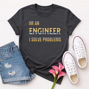 May include: A dark grey short-sleeved t-shirt featuring the text "IM AN ENGINEER I SOLVE PROBLEMS" in a yellow and blue gradient font. The shirt is paired with blue denim shorts, white canvas trainers, and three pink calla lily flowers.