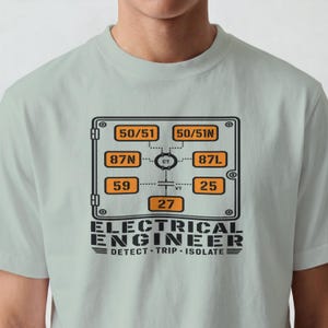 May include: A light sage green t-shirt featuring a black and orange graphic of an electrical relay diagram. The design includes numerical codes like 50/51, 87N, 87L, 59, 25, and 27, with the text "ELECTRICAL ENGINEER" and "DETECT - TRIP - ISOLATE" below.