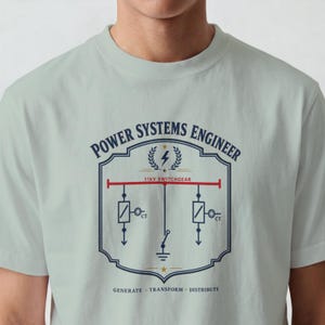 Può includere: Una t-shirt verde salvia chiaro con una grafica blu scuro di uno schema di ingegneria elettrica. Il testo recita POWER SYSTEMS ENGINEER, 11kV SWITCHGEAR e GENERATE TRANSFORM DISTRIBUTE. Il design include un'icona a forma di fulmine.
