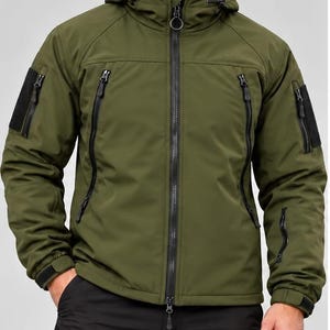 Op de afbeelding: Een olijfgroene tactische softshell jas met een capuchon met trekkoorden, een zwarte ritssluiting over de gehele lengte en meerdere zakken met rits op de borst en mouwen. De mouwen hebben zwarte klittenbandvlakken en verstelbare boorden.