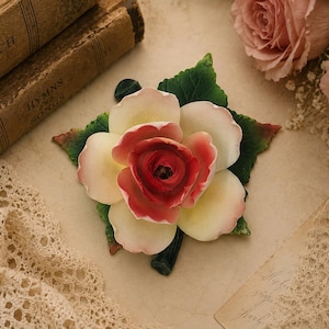Puede incluir: Una figura de rosa de cerámica con pétalos en capas que transicionan desde un centro rojo intenso hasta bordes de color blanco crema. La flor está rodeada por hojas de color verde oscuro texturizadas con venas y un tallo verde.