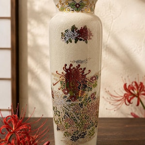 Puede incluir: Un jarrón cilíndrico alto de cerámica con un acabado de esmalte craquelado de color crema. El jarrón presenta intrincados y coloridos motivos florales en rojo, verde y blanco, con detalles en tono dorado alrededor del borde y en los diseños.