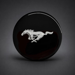 May include: A circular emblem featuring a glossy black finish with a centred, raised silver-toned metallic horse in a galloping pose. The design is smooth and reflective, showcasing a clean, rounded silhouette with a prominent, detailed equine figure.