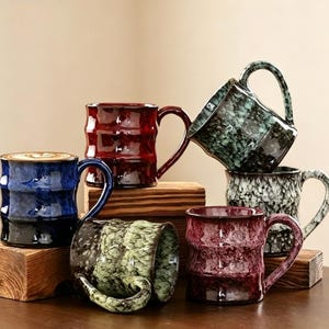 May include: A collection of six ceramic mugs with a textured, bamboo-inspired ridged surface. The mugs feature glossy, multi-coloured reactive glazes in shades of deep blue, dark red, forest green, speckled white, and maroon, each with a curved handle.