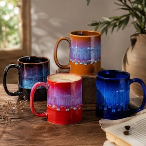 Può includere: Quattro tazze in ceramica con finitura smaltata reattiva. Una tazza è arancione con gocciolature blu, una è nera con toni blu e viola, una è rossa con gocciolature viola e una è blu con un effetto smalto colante. Ogni tazza ha un manico curvo.