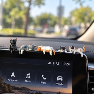 May include: Five miniature cat figurines perched on the top edge of a car infotainment screen. The cats include a solid black cat, a grey tabby, an orange and white cat, a black and white cat, and a calico cat, all in various resting poses.