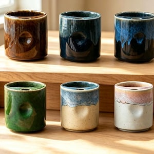 May include: Six cylindrical ceramic cups with a thumb indentation on the side and a small hole on the top rim. The cups feature various reactive glazes in brown, dark blue, green, cream, and pink tones with smooth, glossy finishes.