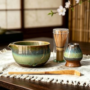 May include: A ceramic matcha bowl with a green and brown drip glaze, a bamboo whisk inside a clear cylindrical case, a ceramic whisk holder with a matching green and brown glaze, and a long, curved bamboo tea scoop on a white fringed cloth.