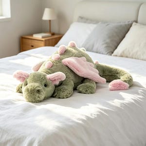 May include: A plush dragon toy featuring soft sage green fur, light pink wings, pink ears, and a pink tail tip. The stuffed animal has a rounded snout, small dark eyes, and a row of small pink spikes running along its back and tail.