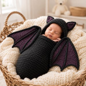 May include: A crochet baby swaddle set featuring a black hooded cocoon with pointed ears and two large, detachable bat wings. The wings and inner ear details are a deep purple color. The swaddle is crafted from dark yarn with a textured stitch.