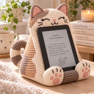 May include: A crocheted cat-shaped tablet or e-reader stand in cream, tan, and brown yarn. The cat features embroidered closed eyes, a pink nose, and pink paw pads. A black e-reader is held in place, displaying the text A quieter kind of happiness.