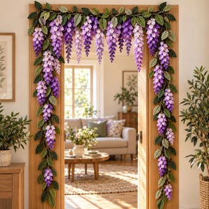 Op de afbeelding: Een decoratieve deurlijstslinger met gehaakte blauweregenbloemen in tinten paars, lavendel en wit, afgewisseld met gestructureerde groene bladeren. Het bloemstuk hangt langs beide zijden en over de bovenkant van het houten deurkozijn naar beneden.