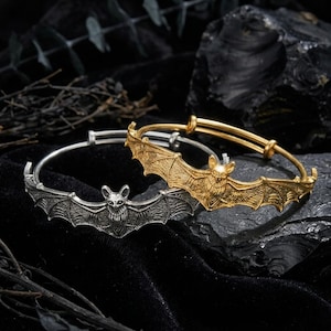 May include: Two adjustable bangle bracelets, one in a silver-tone finish and one in a gold-tone finish. Each bracelet features a central bat motif with detailed wings and textured fur, extending across the front of the circular metal band.