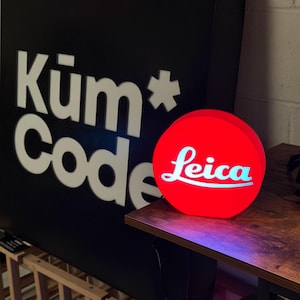 May include: A circular red illuminated sign featuring the white Leica logo in the centre. Behind it stands a large black rectangular sign with the white text Küm* Code displayed in a bold, sans-serif font, creating a high-contrast visual display.
