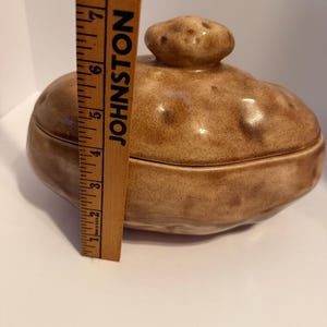 May include: A ceramic lidded container shaped like a potato with a brown, speckled glaze. A wooden ruler is positioned vertically against the container, showing a height of approximately 6 inches from the base to the top of the lid handle.