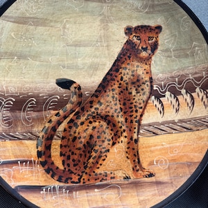 May include: A round decorative plate featuring a painted cheetah in a seated position. The cheetah has a golden-brown coat with black spots. The background consists of horizontal bands in shades of tan and brown with white line art patterns.