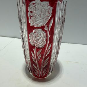 May include: A tall, tapered glass vase featuring a deep red center panel with clear, cut-glass rose floral motifs. The sides of the vase are made of clear, textured glass with a vertical, wavy pattern that creates a scalloped, uneven top rim.