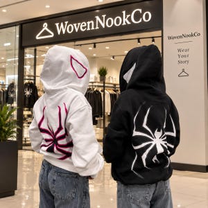 Op de afbeelding: Een witte hoodie met een roze en zwarte spinnengrafiek op de achterkant en een roze spinoogdetail op de capuchon, gecombineerd met een zwarte hoodie met een grote witte spinnengrafiek op de achterkant en witte spinoogdetails op de capuchon.