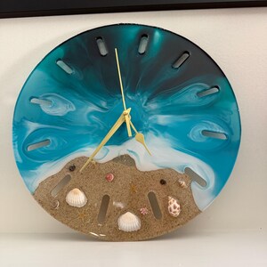 May include: A round wall clock featuring a resin design of a beach with turquoise and deep blue water swirling into white foam waves. The lower section contains sand embedded with various small seashells and starfish, with gold clock hands.