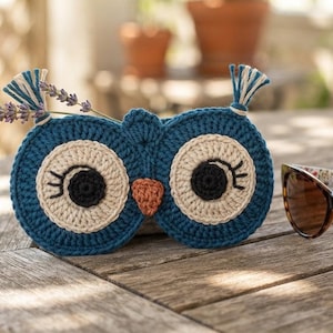 May include: A blue crocheted owl-shaped pouch featuring large cream-colored eye patches, black pupils with stitched eyelashes, and a small orange beak. Two small tassels sit atop the owl's head, with a sprig of lavender tucked behind one.
