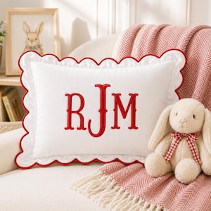 May include: A rectangular white throw pillow featuring a scalloped edge trimmed in red piping. The center of the pillow displays the embroidered red monogram letters RJM in a classic serif font. The pillow is positioned next to a plush toy rabbit.