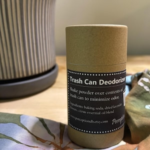 May include: A cylindrical cardboard container of trash can deodorizer with a black label featuring white text that reads Trash Can Deodorizer, Shake powder over contents of trash can to minimize odor, ingredients, and a website address.