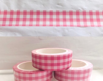May include: Three rolls of pink and white checkered washi tape are stacked on top of each other. A strip of the same tape is shown above the rolls. The tape has a gingham pattern and is likely used for crafting or decorating.