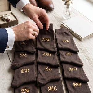 May include: A collection of dark brown socks arranged in three columns, each featuring gold embroidered initials including AG, HL, ED, TM, DM, LL, ML, DL, MO, and HB. The socks are laid out flat, showcasing the custom monogramming on each individual pair.
