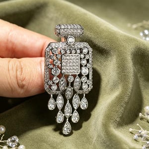 Puede incluir: Un broche de tono plateado con forma de frasco de perfume, incrustado con numerosos diamantes de imitación transparentes y brillantes. El diseño presenta un cuerpo rectangular con un panel central cuadrado y flecos colgantes de cristales en forma de lágrima.