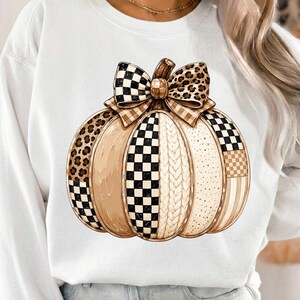 Op de afbeelding: Een wit sweatshirt met lange mouwen en een grote centrale pompoenafbeelding. De pompoen is verdeeld in verticale segmenten met gevarieerde patronen, waaronder zwart-wit geblokte motieven, bruine luipaardprint en crèmekleurige kabelbreistructuren.