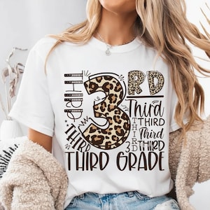 May include: A white short-sleeve t-shirt featuring a large graphic design with a leopard print number 3, the text 3RD, and the word THIRD repeated in various fonts and sizes, culminating in the bold text THIRD GRADE at the bottom of the print.