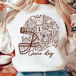 Puede incluir: Una sudadera blanca de cuello redondo con un gran gráfico marrón en forma de casco de fútbol americano. El diseño incluye varios iconos relacionados con el fútbol, como un estadio, una camiseta, un marcador y el texto Game day en cursiva.