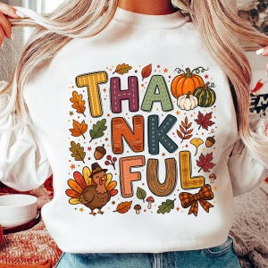 May include: A white crewneck sweatshirt featuring a colourful graphic with the text THANKFUL. The design includes a turkey, pumpkins, acorns, mushrooms, and autumn leaves in shades of orange, yellow, green, brown, and red, with a plaid bow.